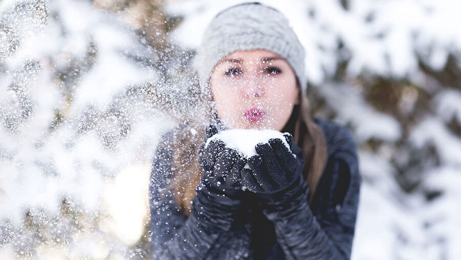 junge Frau mit grauer Mütze in Winterlandschaft pustet Schnee, den sie in Händen hält