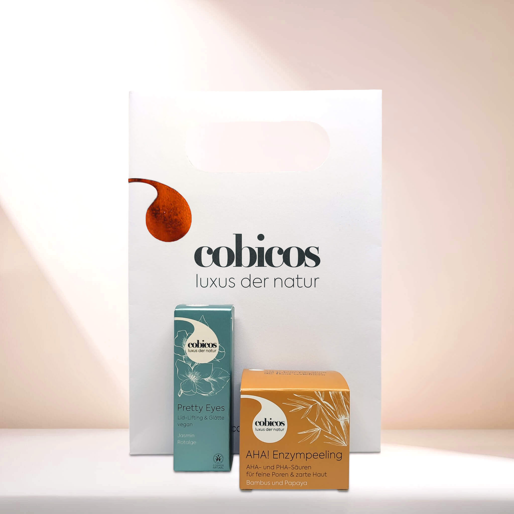 weiße cobicos Papiertragetasche mit Logo. Davor steht die Pretty Eyes in türkisfarbener Verpackung und das AHA! Enzympeeling in leuchtend oranger Verpackung